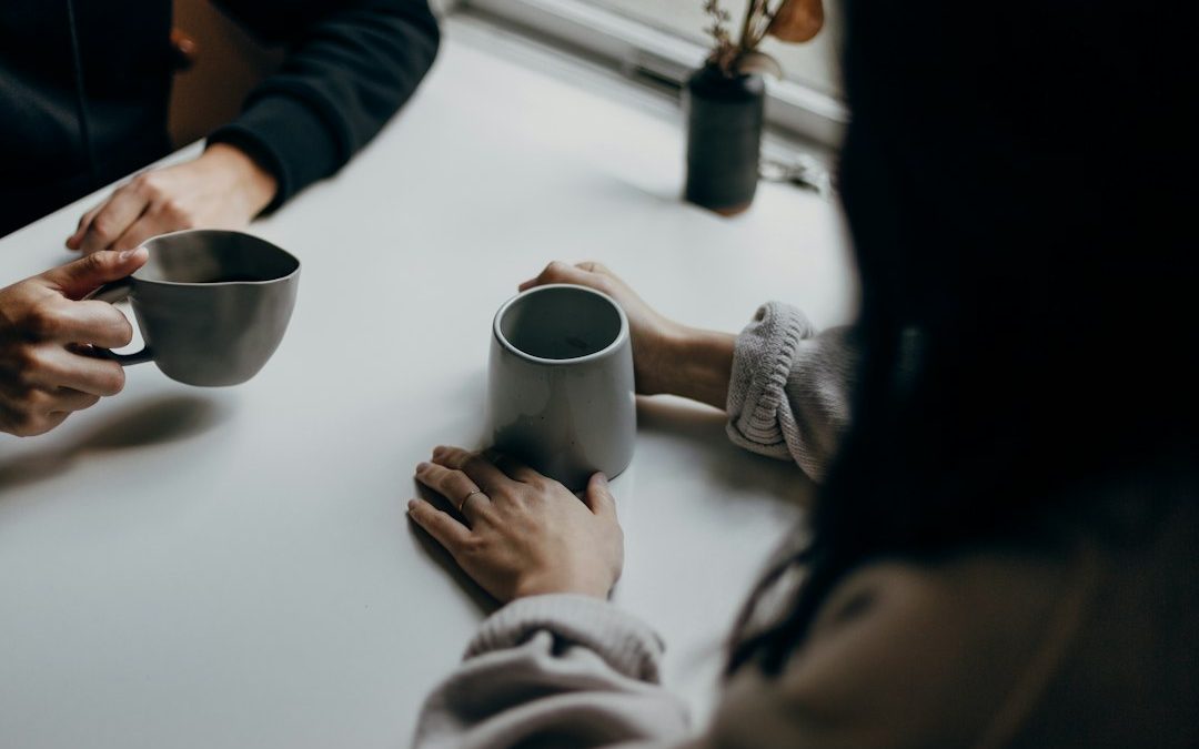 Two people holding coffee mugs across a table in conversation
